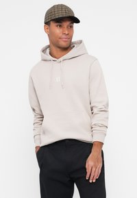 Felpa con cappuccio color beige chiaro con cordini e un piccolo logo "II" bianco sul petto, abbinata a pantaloni neri e un berretto stampato.