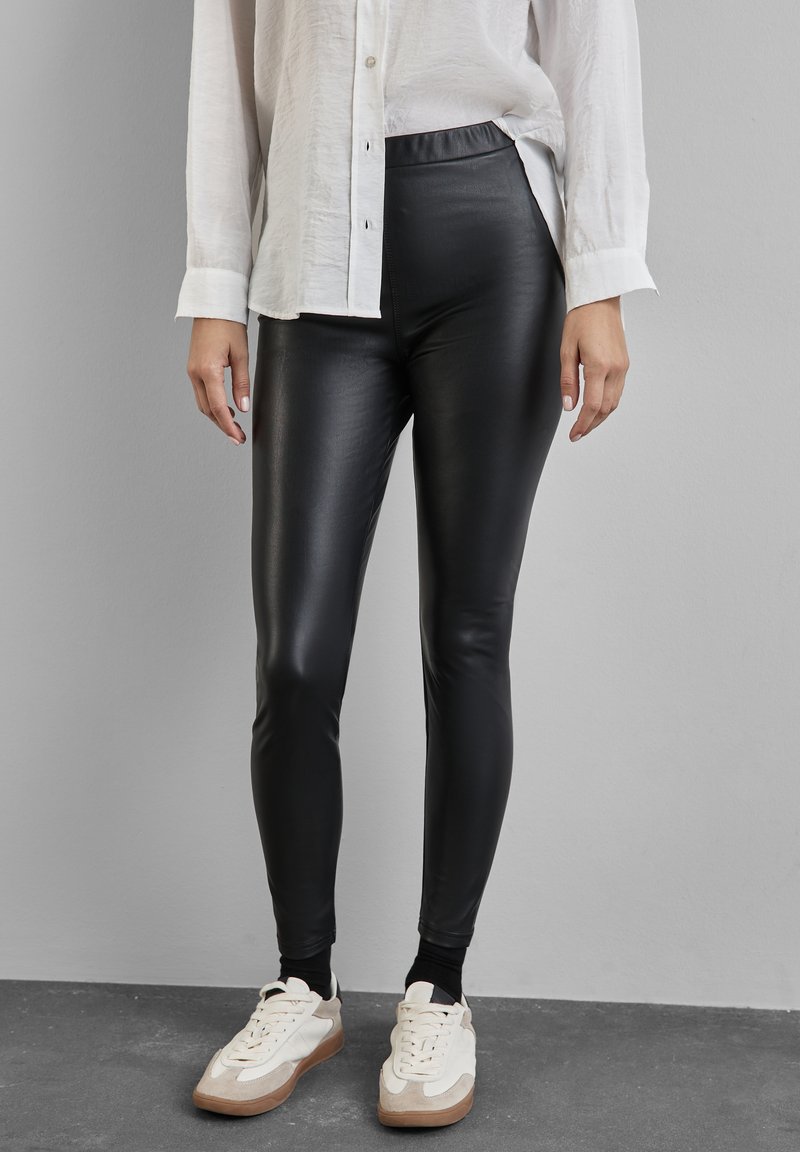 Leggings neri in ecopelle con una texture liscia, vita alta elasticizzata, design aderente, abbinati a una camicia bianca con bottoni e scarpe sportive.
