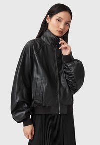 Černá kožená bomber bunda s vysokým límcem, elastickými manžetami a bočními kapsami. Vyznačuje se hladkou texturou a uvolněným střihem.
