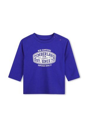 Chemise bleue à manches longues pour tout-petit avec logo Timberland blanc et texte, avec deux boutons-pression aux épaules facilitant l'habillage.