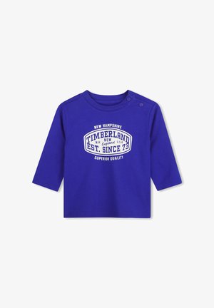 Chemise bleue à manches longues pour tout-petit avec logo Timberland blanc et texte, avec deux boutons-pression aux épaules facilitant l'habillage.