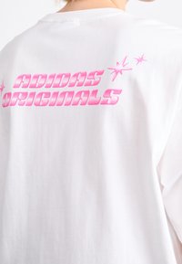 T-shirt bianco in cotone con logo rosa "ADIDAS ORIGINALS" e accenti a forma di stella. Il design è centrato sulla schiena.