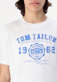 Balta medvilninė marškinėliai su mėlynu grafiniu tekstu, kuriame yra "TOM TAILOR," "1962," ir "GENERAL PURPOSE UTILITY." Apvalus iškirpimas, trumpomis rankovėmis.