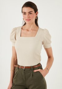 Top beige à côtes avec des manches courtes bouffantes et un décolleté carré, associé à un pantalon vert et une ceinture marron. Tissu lisse avec une texture subtile.