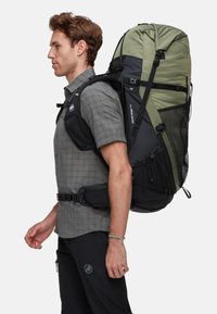 Mochila grande, negra y verde, con bolsillos laterales de malla y correas ajustables, llevada por una persona con una camisa gris a cuadros y pantalones oscuros.