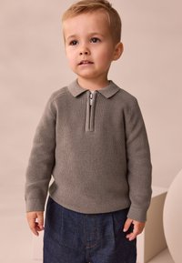 Grauer Strickpullover mit Reißverschlusskragen, langen Ärmeln und strukturiertem Rippstrick. Kombiniert mit dunklen Jeansshorts, die große Taschen haben.