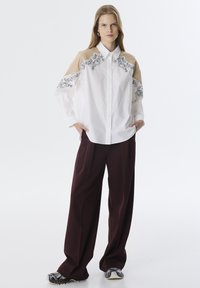 Camisa blanca de botones con tejido beige en los hombros, con bordado floral negro. Combinada con pantalones holgados color burdeos y zapatillas con estampado.