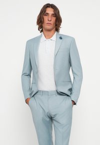 Costume bleu clair à coupe ajustée, fermeture à un bouton et détail de poche, porté sur un polo blanc à motif texturé.