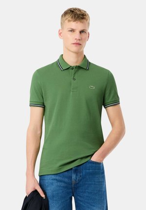 Uomo biondo che indossa una polo verde con bordi bianchi e neri e jeans blu, tiene una giacca scura in una mano.