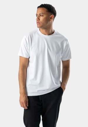 Hvid t-shirt med korte ærmer og rund halsudskæring, lavet af et glat stof. Modellen bærer den med afslappede sorte bukser.
