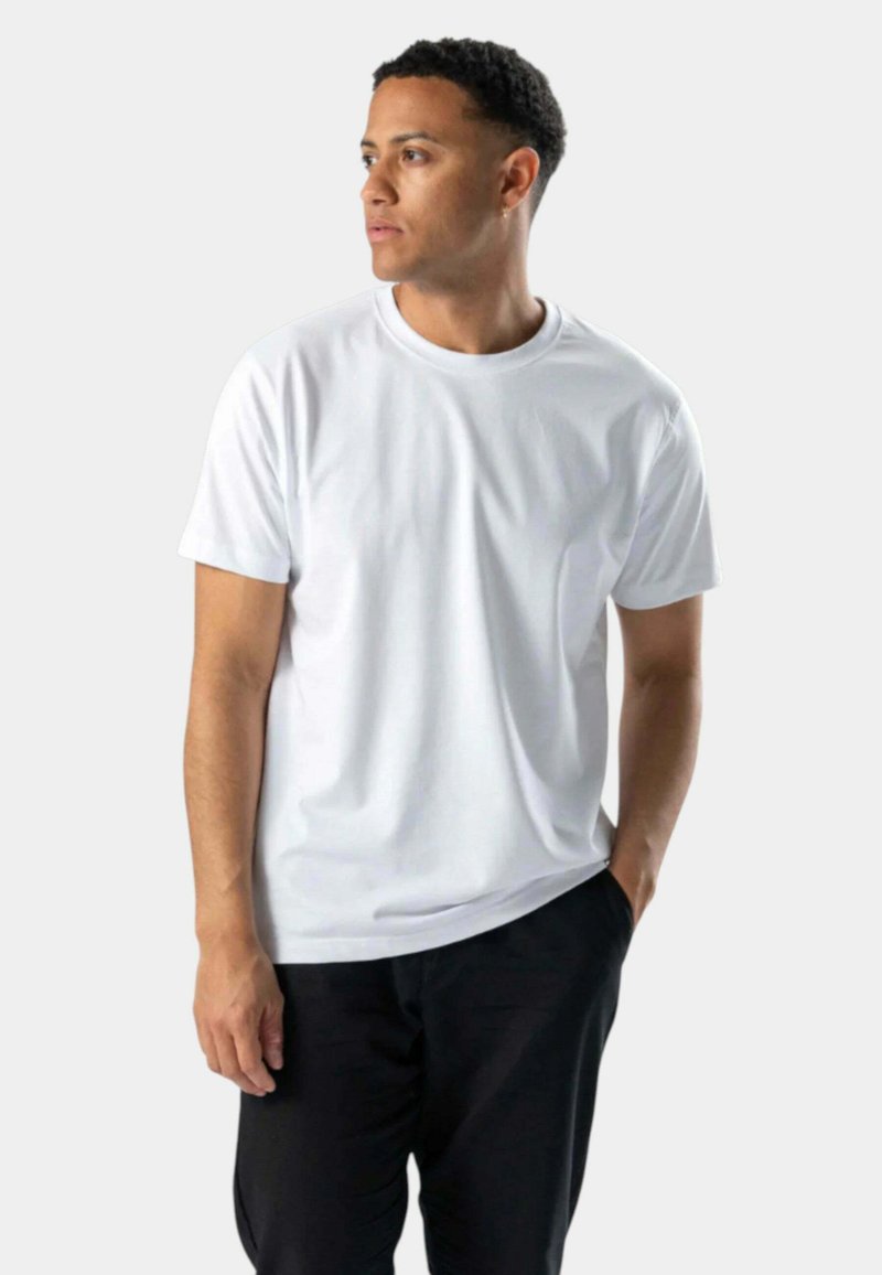 Hvid t-shirt med korte ærmer og rund halsudskæring, lavet af et glat stof. Modellen bærer den med afslappede sorte bukser.