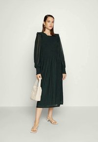 Vestido verde-escuro de mangas longas com tecido texturizado, decote com babados e cintura franzida. Bolsa branca e sandálias com tiras completam o visual.