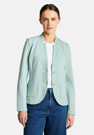 Frau trägt ein hellgrünes Blazer über einem weißen Hemd und blauen Jeans, stehend vor einem schlichten weißen Hintergrund.
