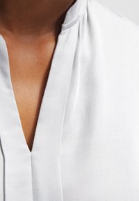 Blusa bianca con scollatura a V, realizzata in tessuto liscio. Il design include texture sottili e dettagli di cucitura lungo la scollatura.
