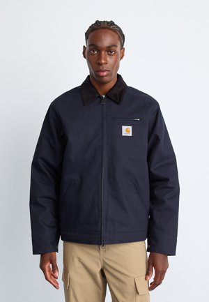 Giacca blu navy realizzata in tessuto resistente, con colletto marrone e patch con logo, chiusura con zip e tasche laterali.