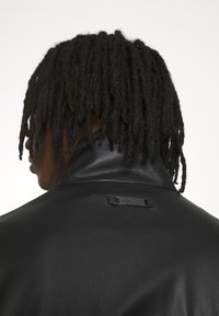 Svart läderjacka med hög krage, som har en liten märkesetikett på baksidan. Slät textur med minimala utsmyckningar.