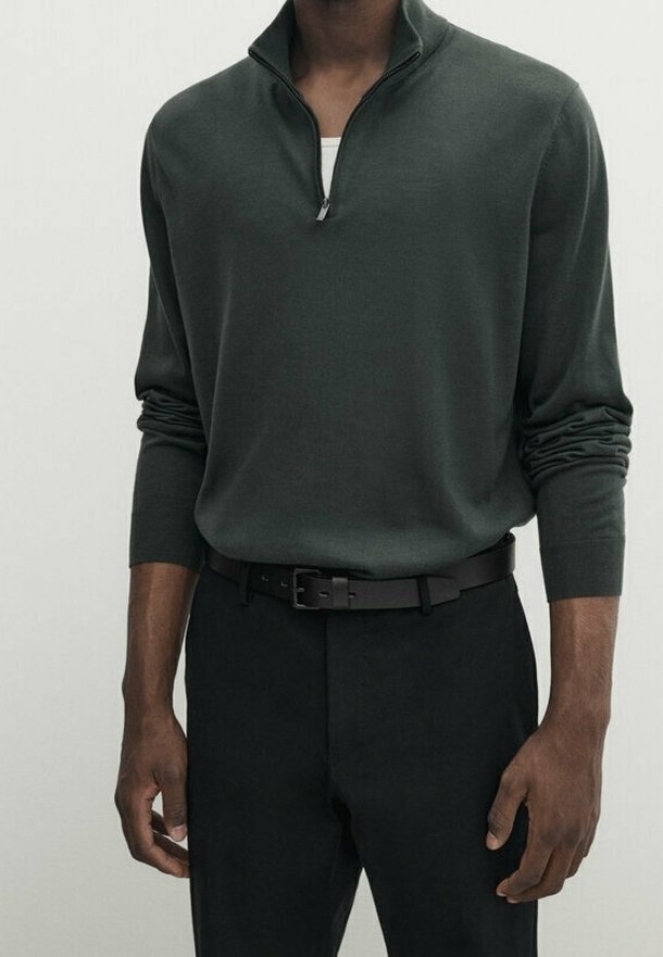Pull à col zippé vert foncé en tissu doux, avec manches longues et poignets détendus ; associé à un pantalon noir et une ceinture.