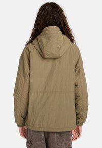 Persoon met lang, krullend haar, gekleed in een lichtbruine hooded jas en donkere cargobroek, gezien van achteren tegen een witte achtergrond.