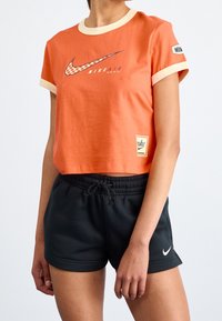 Oranje cropped T-shirt met crème accenten en Nike-logo. Gecombineerd met zwarte shorts met trekkoord en een kleine witte Nike-swoosh.