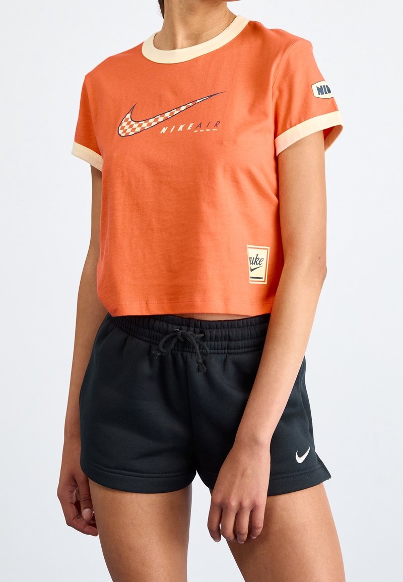 Oranje cropped T-shirt met crème accenten en Nike-logo. Gecombineerd met zwarte shorts met trekkoord en een kleine witte Nike-swoosh.