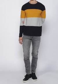 Randig stickad tröja i svart, senapsgul, grå och marinblå med rund halsringning. Bärs med grå jeans och svarta sneakers.