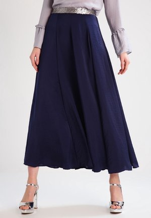 Kobieta w granatowej spódnicy za kolano z plisami, z srebrnym paskiem wysadzanym cekinami, jasnoszara bluzka z falbankami przy mankietach oraz srebrne sandały na wysokim obcasie.