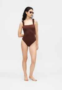 Femme portant un maillot de bain une pièce marron foncé texturé avec des bretelles froncées et de petites lunettes de soleil ovales, debout pieds nus sur un sol blanc.