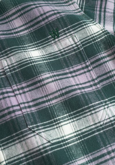 Tissu en tartan vert et lavande avec une texture douce. Présente un petit logo brodé et de grandes lignes horizontales et verticales.