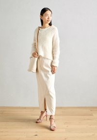 Pull en tricot beige clair, jupe longue crème avec une fente, talons roses à lanières et sac en osier. Mannequin se tient sur un sol en bois contre un mur neutre.