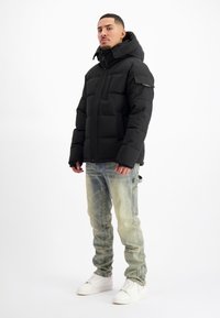 Zwarte pufferjas met hoge kraag, voorzien van een ritssluiting aan de voorkant en zijzakken. Gemaakt met lichtblauwe denim jeans en witte sneakers.