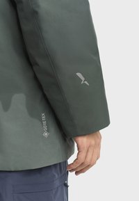 Zelená Gore-Tex bunda s hladkým textúrovým rukávom. Obsahuje šedé logo blízko manžety a reflexný detail.