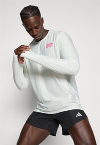Sportler passt das Daumenloch am langärmligen BMW Berlin Marathon Shirt an, trägt schwarze Adidas Laufshorts vor einem schlichten Hintergrund.
