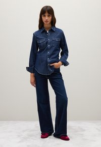 Woman wearing dark blue denim shirt and wide-leg jeans, standing with one hand in pocket and red shoes on light carpet.
