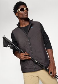 Chaleco aislante negro con cremallera, un logo azul y detalle de malla; camiseta de manga larga negra; gafas de sol beige; bastón de trekking negro.