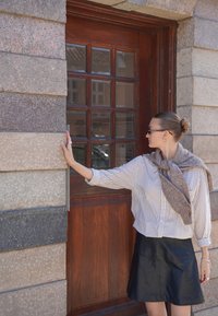 Csíkos ing, sötét bőrszoknya, szürke sál, egy négyzet alakú üvegablakokkal ellátott faajtó mellett áll, texturált kőfal háttérrel.