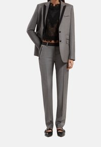 Grijze op maat gemaakte kostuum met een gestructureerde blazer, bijpassende broek en een doorschijnend zwart kanttop. Zwarte sandalen met straps maken de outfit compleet.