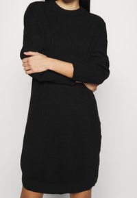 Vestito a maglia nero a coste con maniche lunghe, scollo rotondo e tasche laterali, caratterizzato da una texture orizzontale su tutta la superficie.