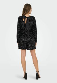 Sort paillet romper med lange ærmer, åben ryg og stor sort sløjfe. Tekstureret, skinnende materiale med tætsiddende shorts og ankelremssko.