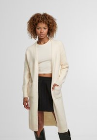 Dlouhý, lehký cardigan v krémové barvě, s volným střihem, dvěma předními kapsami a žebrovanými manžetami. Nositý přes zkrácený top a sukni.