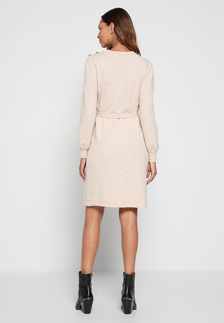 Robe tricotée beige à longueur genou avec des manches longues, une ceinture à nouer à la taille, et des accents côtelés à l'ourlet, associée à des bottines noires.