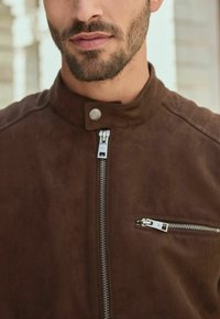 Brūna zamšāda jaka ar augstu apkakli, ar sudraba priekšējo rāvējslēdzēju un papildu rāvējslēdzēju krūšu kabatu. Gluda tekstūra, minimālistisks dizains.