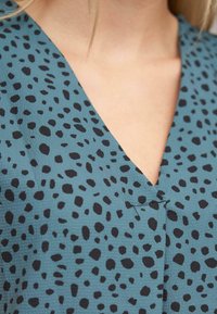 Close-up van een blauw-groen kledingstuk met zwarte onregelmatige vlekken, met een V-halslijn en een zichtbare losse zwarte draad.