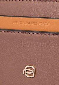 Bruine leren oppervlak met een gestikte oranje strook bedrukt met "Piquadro" en een klein zilverkleurig metalen logo eronder.