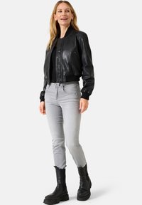 Giacca bomber in pelle nera con polsini a costine, abbinata a jeans slim-fit grigi chiari e stivali neri robusti. Design minimalista, senza motivi visibili.
