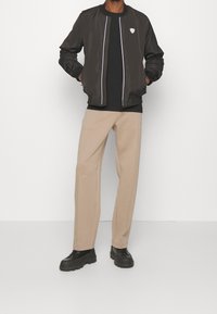 Svart bomberjacka med vit dragkedja, bärs över en svart t-shirt, tillsammans med beige vida byxor och svarta klumpiga skor.