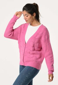 Roze gebreide cardigan met een gestructureerd patroon, v-hals en knoopsluiting. Geribbelde mouwen en zoom, gecombineerd met een witte top en blauwe jeans.