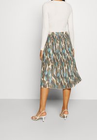 Jupe midi plissée avec un motif abstrait multicolore dans des teintes de beige, bleu et vert. Associée à un haut ajusté à manches longues blanc et des talons blancs.