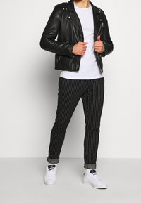 Czarna skórzana kurtka motocyklowa na białym t-shircie, zestawiona z czarnymi spodniami w prążki i białymi trampkami. Widoczne zamki błyskawiczne i mankiety.