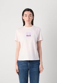 T-shirt di cotone rosa chiaro con maniche corte e scollatura rotonda. Presenta una grafica quadrata viola con "cK" in bianco. Indossata con jeans blu scuro.