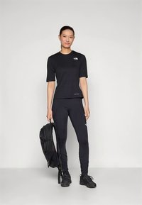 Μαύρο αθλητικό μπλουζάκι και κολάν με λογότυπα. Το μοντέλο κρατάει ένα μαύρο σακίδιο, φορώντας μαύρα αθλητικά παπούτσια. Απλό φόντο.